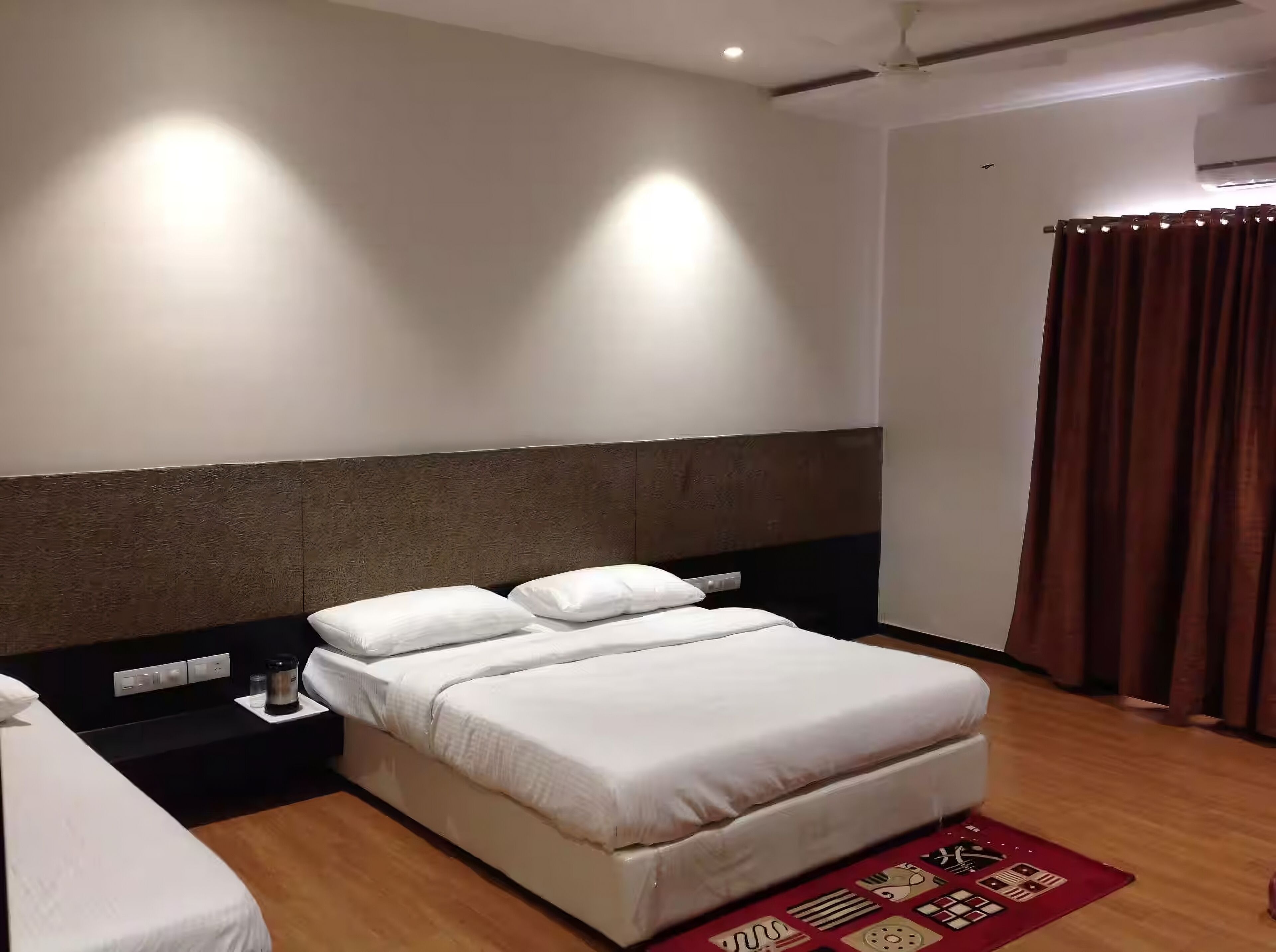 Una habitación de hotel con una cama grande, una mesita de noche, un tapete rojo estampado y una ventana con cortinas.