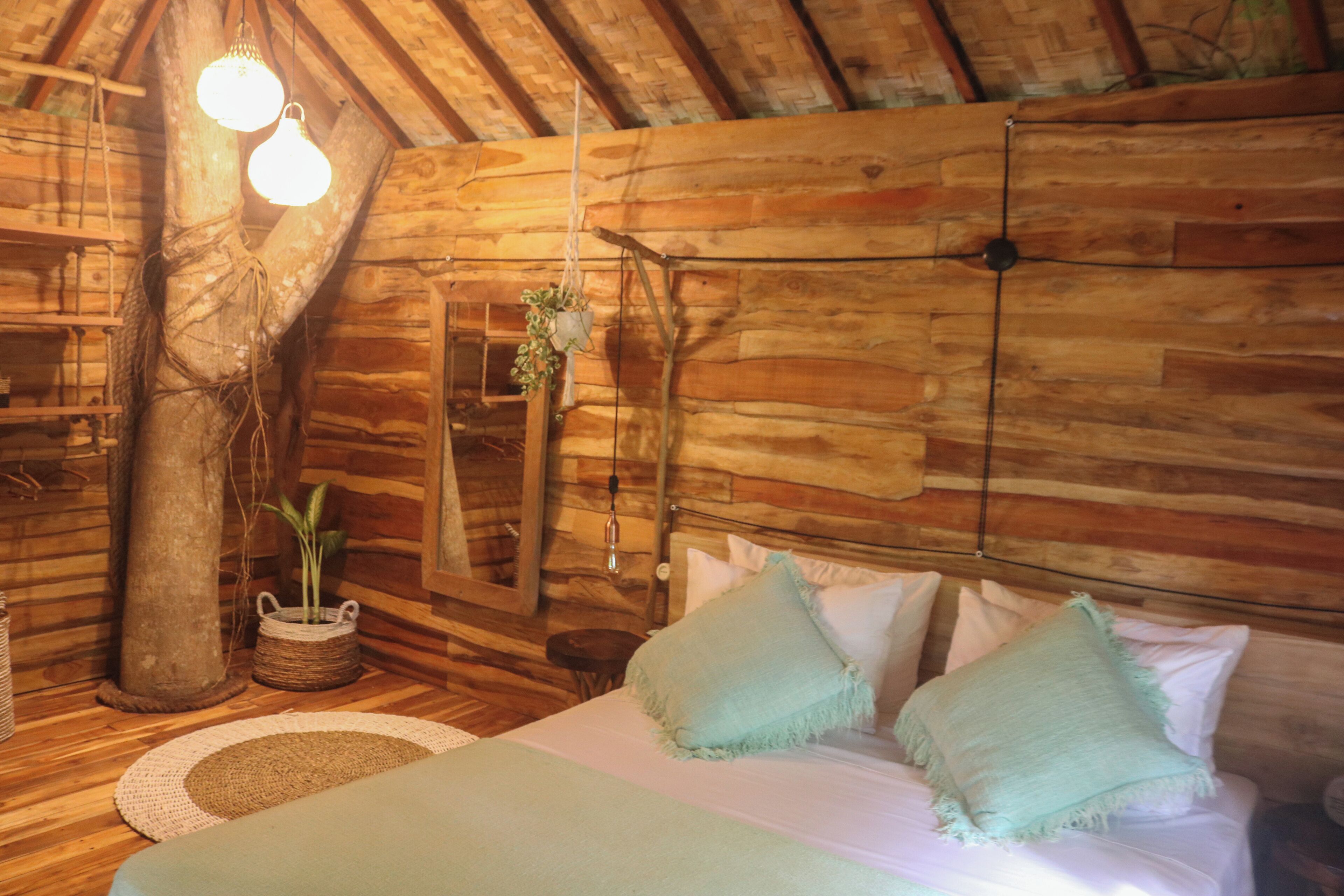 A rustic wooden room with a bed, hanging light fixtures, and a woven basket with plants.