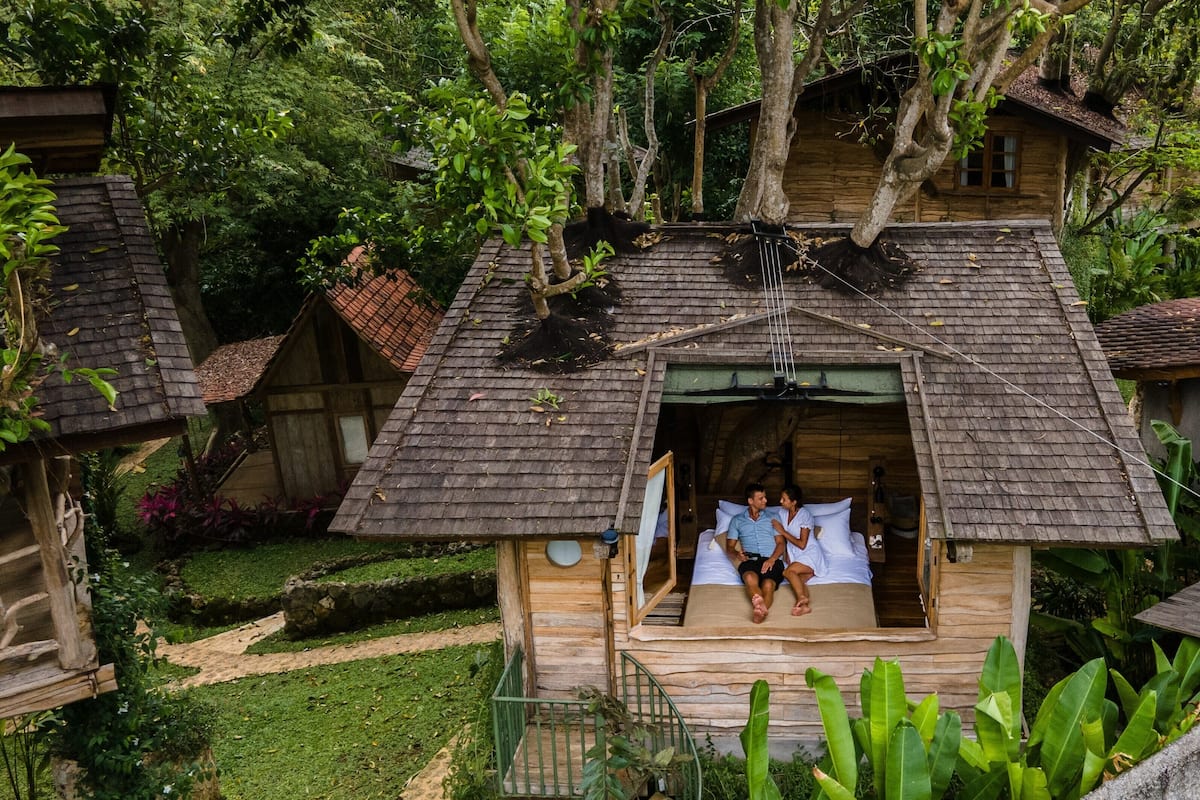 A rustic wooden cabin with a thatched roof, a small porch, and a bed with a white mattress and pillows.