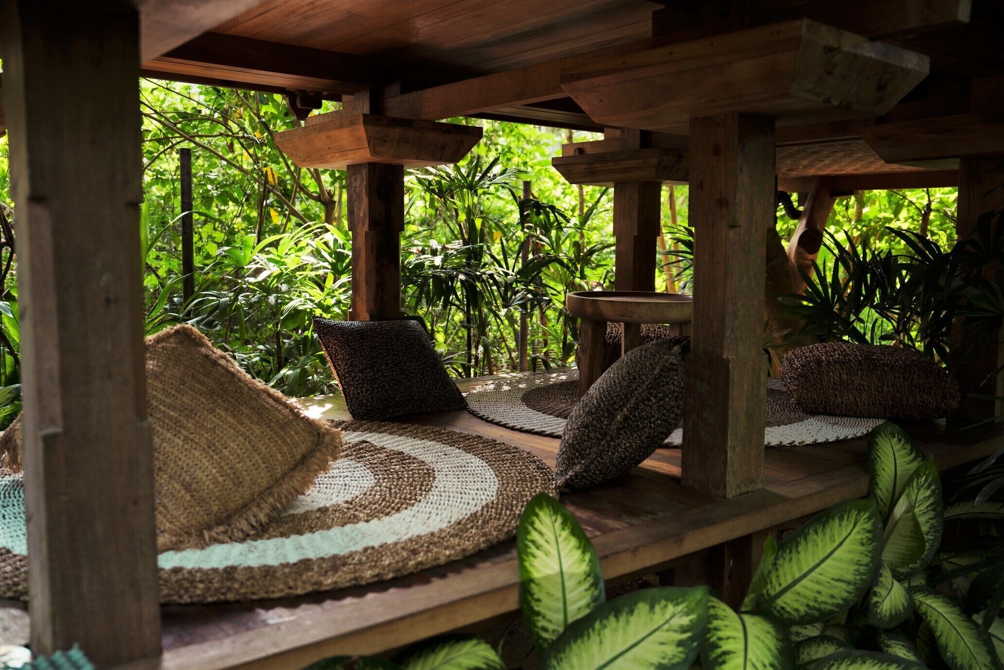A wooden deck with woven mats and pillows, surrounded by lush greenery.