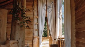 A rustic wooden cabin interior with a wooden bench, hanging plants, and a window with curtains. - Roots Tree House (Ungasan)