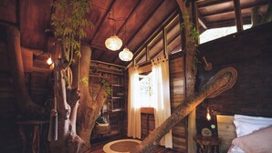 A rustic wooden cabin interior with a large tree trunk as a central feature, a bed, a wooden shelf, and a hanging light fixture. - Roots Tree House (Ungasan)