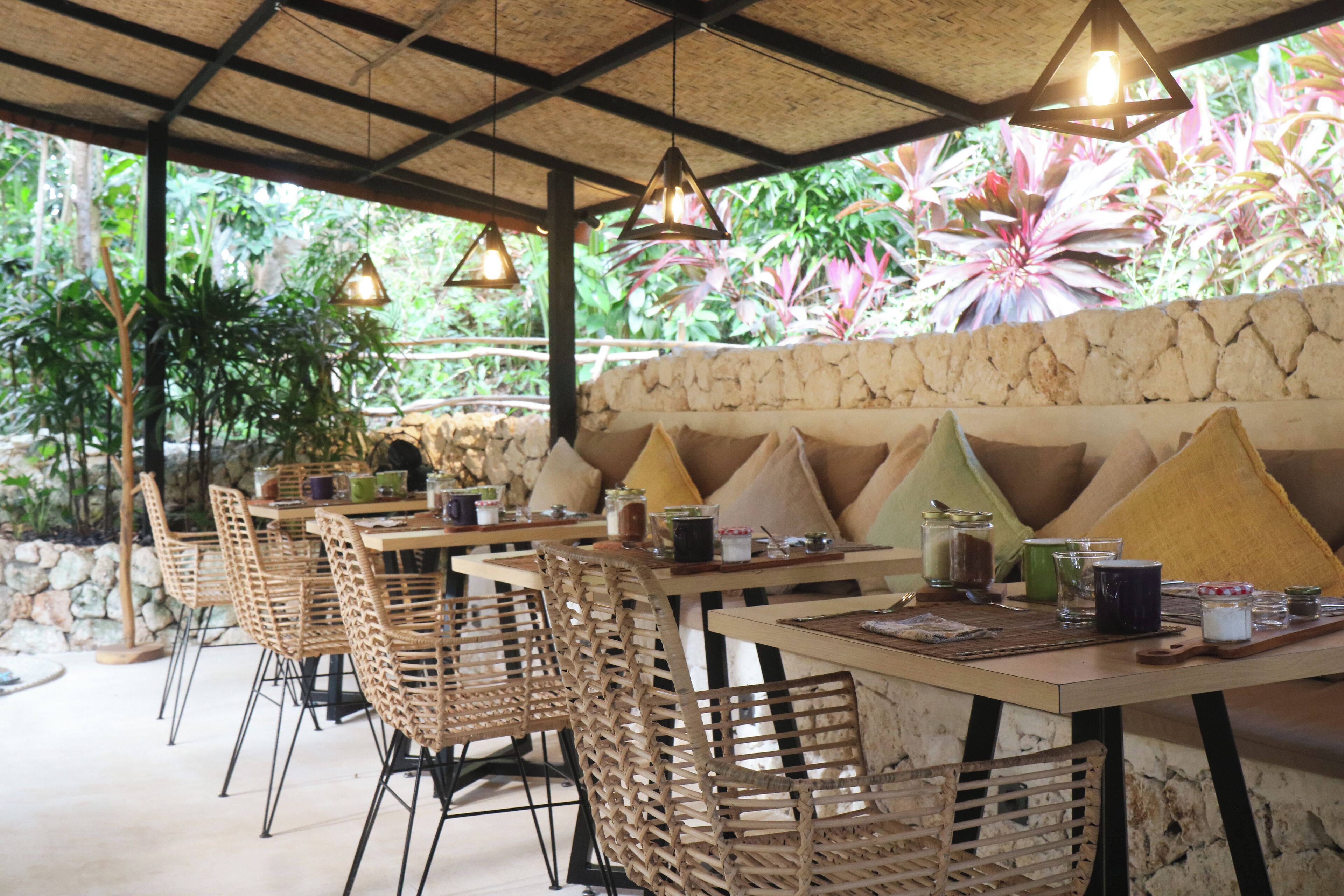 Un espace de restauration en plein air avec des chaises et des tables en osier, des murs en pierre et des lampes suspendues.