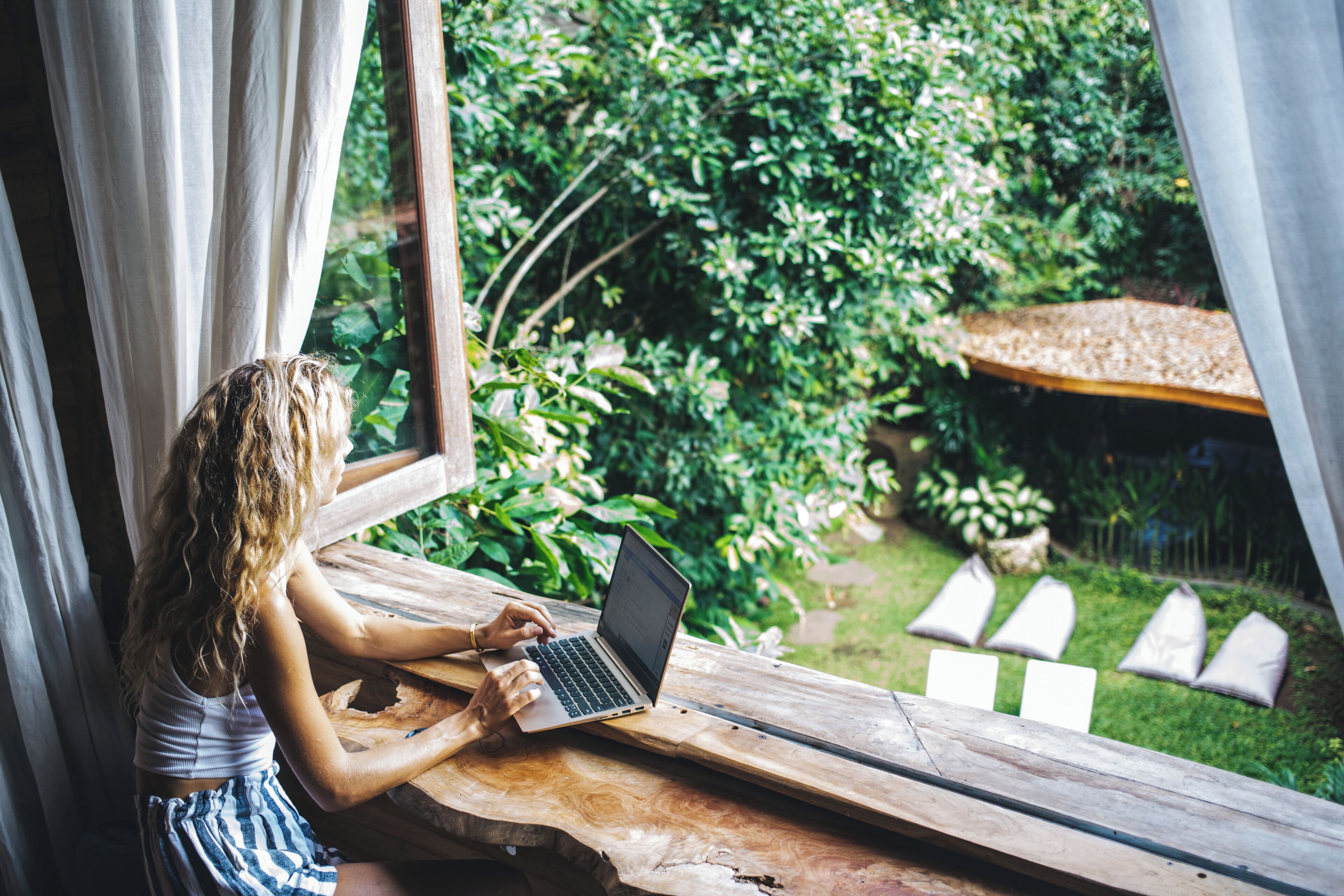 A person using a laptop at a wooden table by a window with curtains, overlooking a garden with greenery and a wooden deck.