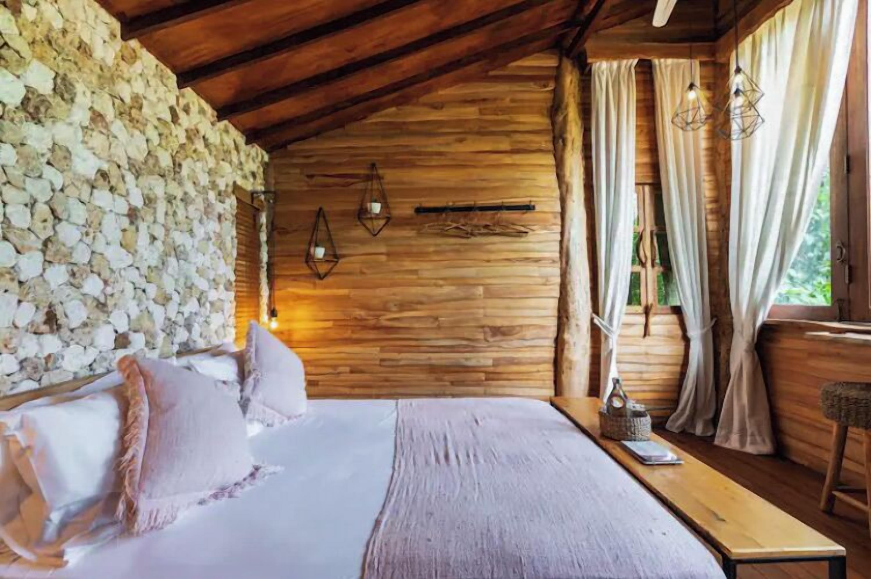 Una habitación de cabaña de madera con pared de piedra, una cama con almohadas, un banco y lámparas colgantes.