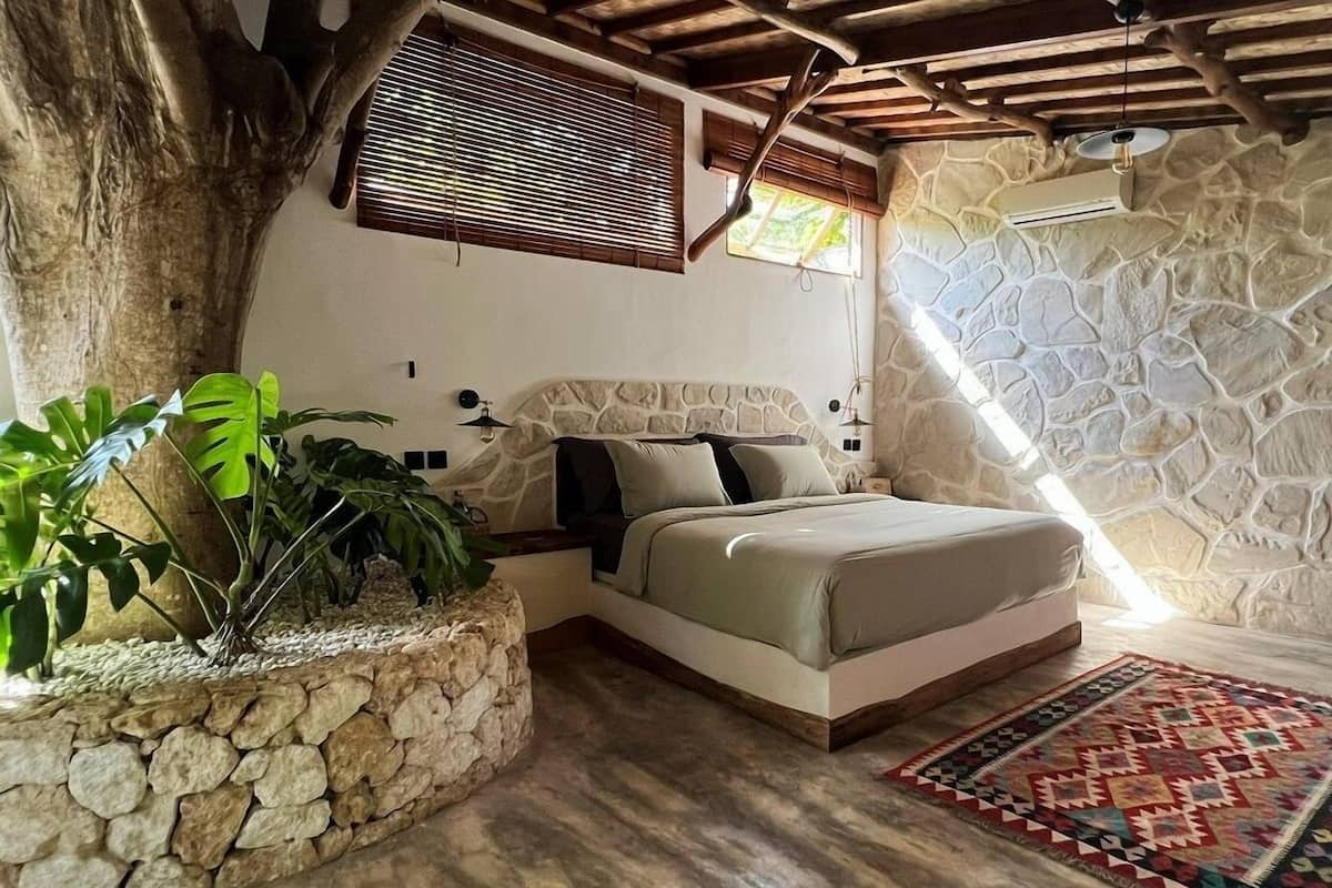 A bedroom with a stone wall, a bed with a stone headboard, a wooden ceiling, and a large window with blinds.