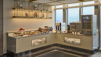 A modern hotel lobby with a marble reception desk, a coffee machine, a beverage dispenser, and a window with a view.