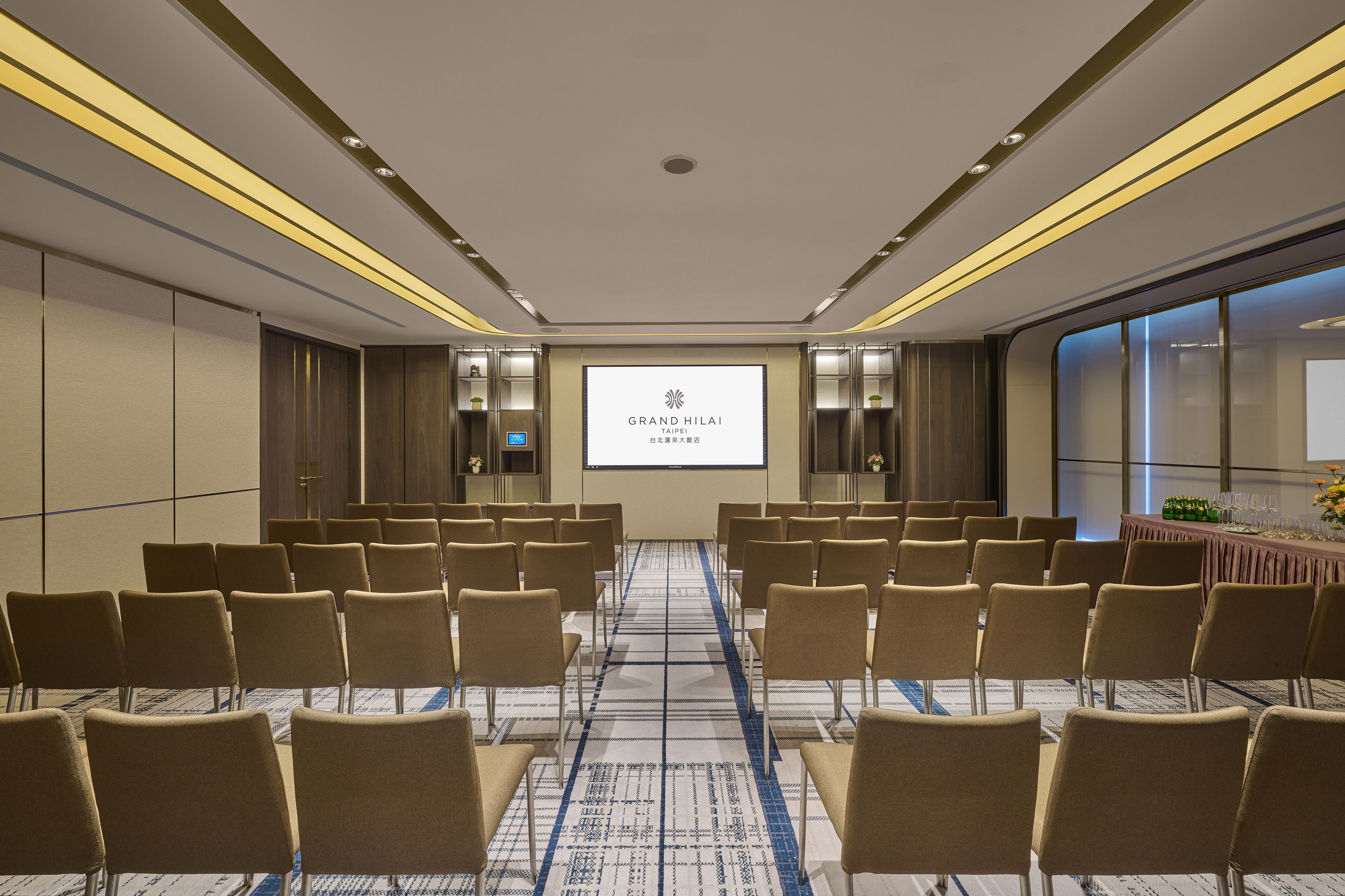 Un salón de conferencias con filas de sillas, una pantalla de proyección mostrando 'GRAND HILAI TAIPEI' y piso alfombrado.