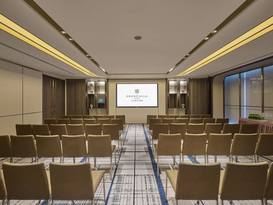 Un salón de conferencias con filas de sillas, una pantalla de proyección mostrando 'GRAND HILAI TAIPEI' y piso alfombrado.