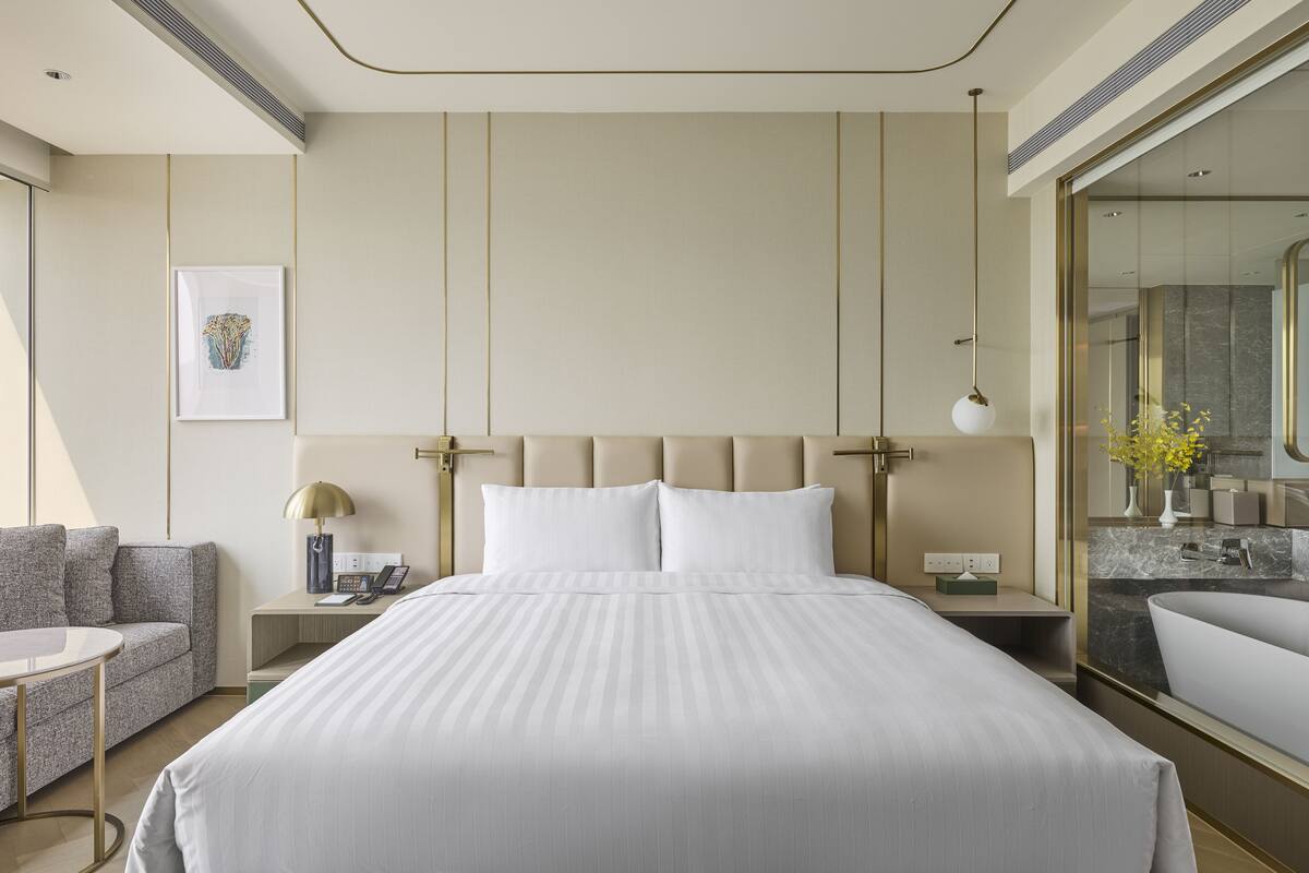 A modern hotel room with a large bed, a sofa, a bedside table with a lamp, and a bathroom visible through a glass partition.