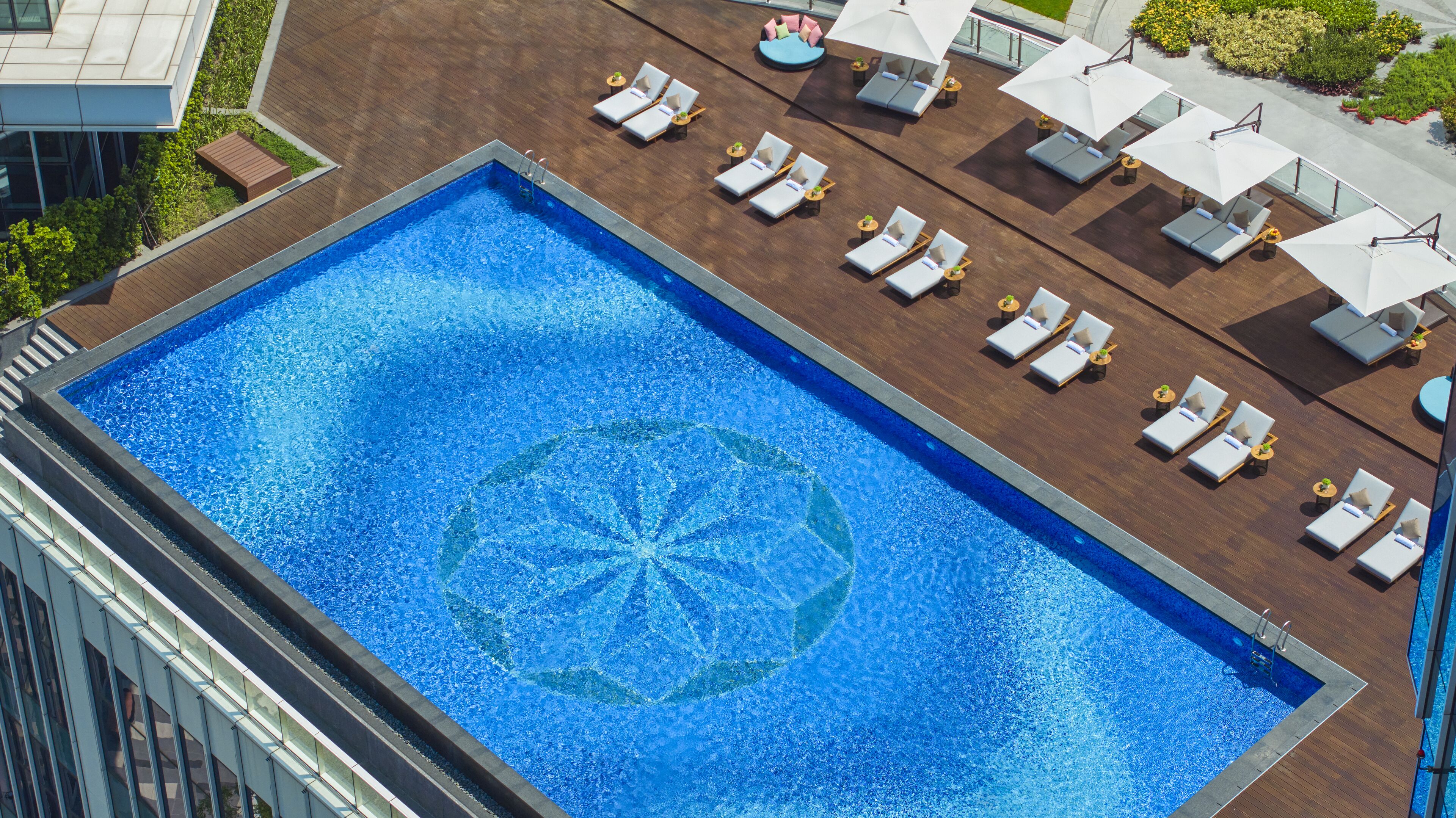 A rooftop pool with a geometric pattern, surrounded by sun loungers and umbrellas, with a modern building in the background.