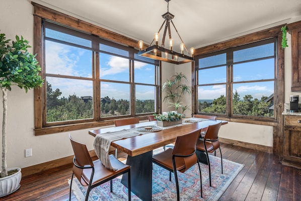 A dining area with a wooden table, chairs, and a chandelier. Large windows offer a view of trees and a clear sky.