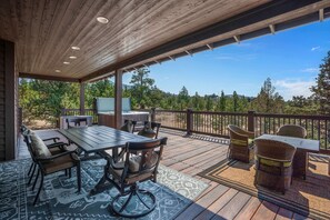 A covered outdoor patio with a wooden table and chairs, a TV, and a scenic view of trees and hills.