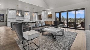 A spacious living area with a grey sofa, a coffee table, and a dining table. There is a kitchen with white cabinets and a large window. - Brasada Ranch | High Desert Modern Luxury | Sauna (Powell Butte)