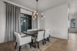 Een eetruimte met een donker houten tafel, witte stoelen, een kamerkleed met een patroon en een moderne kroonluchter.
