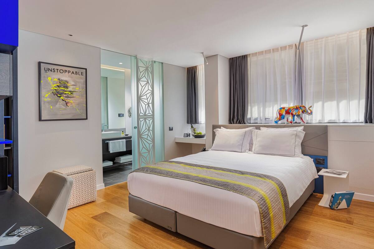 A modern hotel room with a large bed, a desk, and a bathroom visible through a glass door.