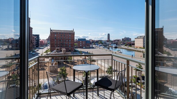 Un balcón con dos sillas y una mesa, con vista a la orilla del agua, edificios y un canal.