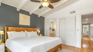 Camera da letto con un letto in legno, biancheria bianca, una ventola a soffitto e vista sul bagno.