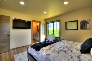 A bedroom with a large bed, a television mounted on the wall, a window with a view of greenery, and a door leading to a bathroom. - Summit Sky Retreat, Red Lodge (Red Lodge)