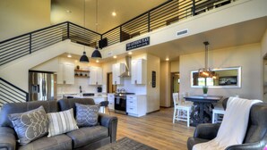 A spacious living area with a sofa, armchair, and dining table. A kitchen is visible in the background with a sign reading "The Gathering Place."