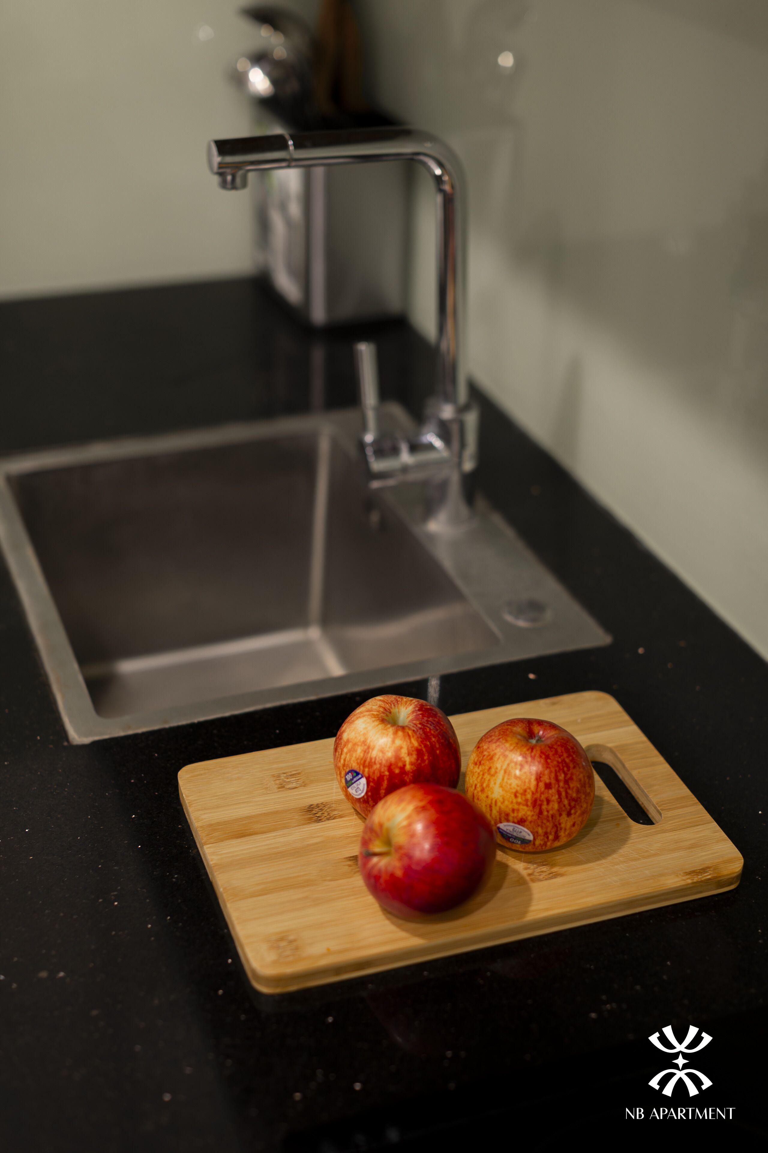 Une cuisine avec un évier en acier inoxydable, une planche à découper en bois sur laquelle reposent deux pommes, et le logo de NB Apartment.