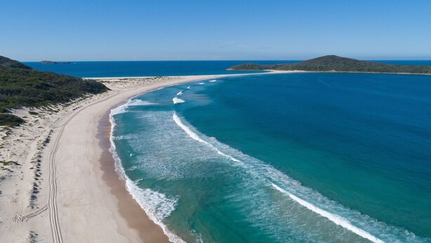 Vista aérea de una playa con aguas azules cristalinas, orillas de arena y un paisaje montañoso al fondo.