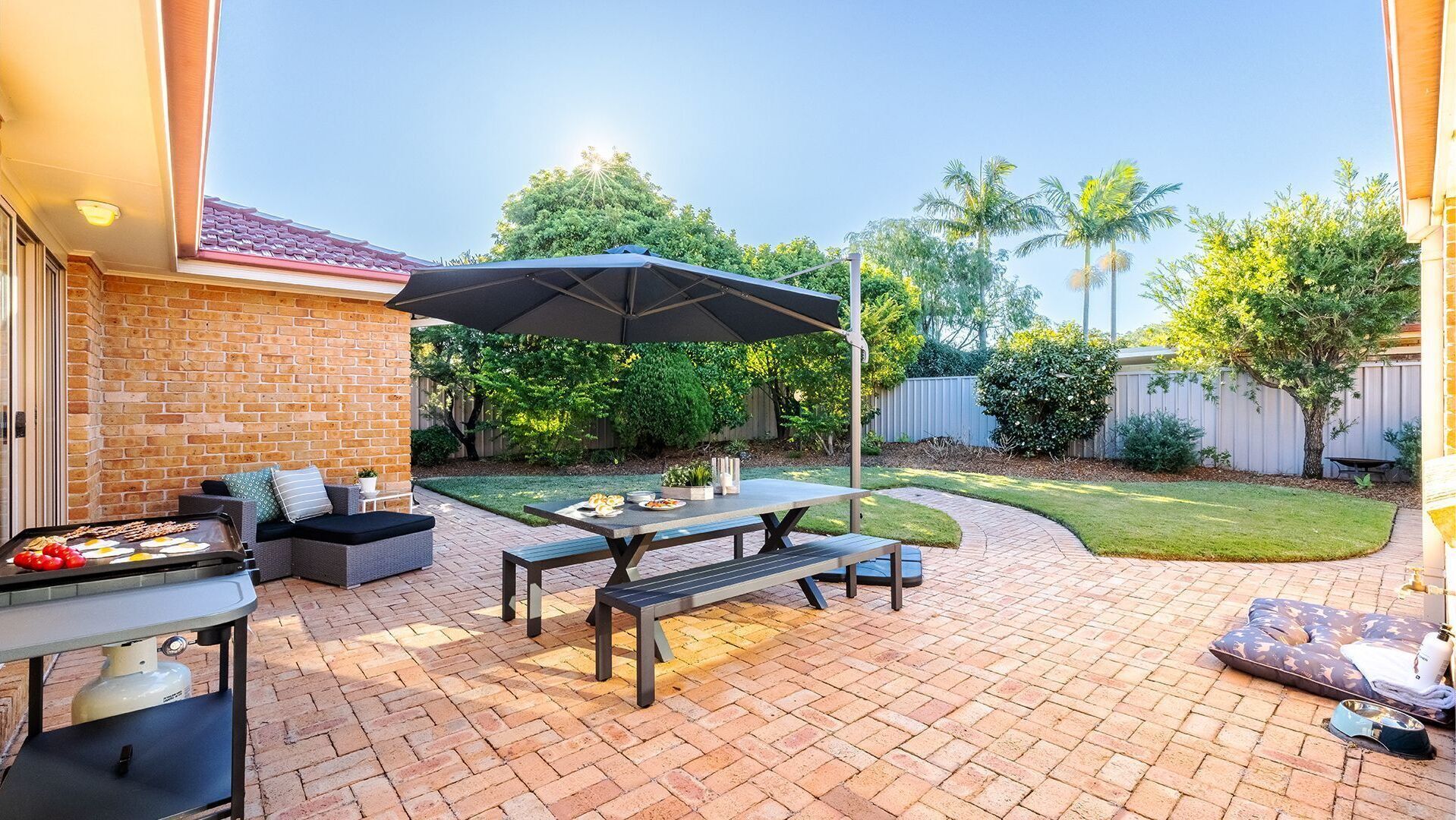 A backyard patio with a brick floor, a table with chairs, a grill, and a covered seating area.