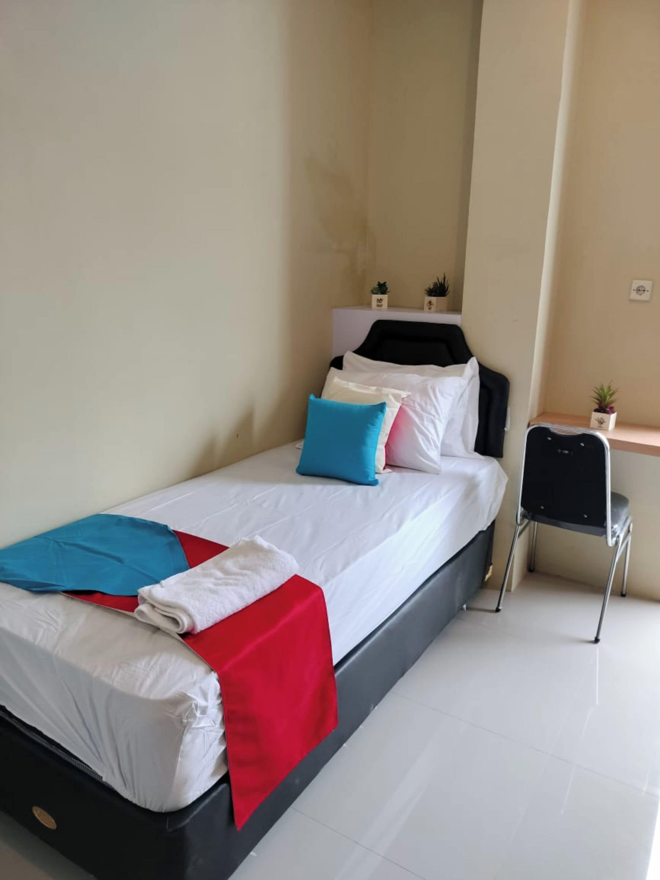 A single bed with white bedding, a red blanket, and blue and pink pillows. There is a small table with a plant and a chair beside the bed.