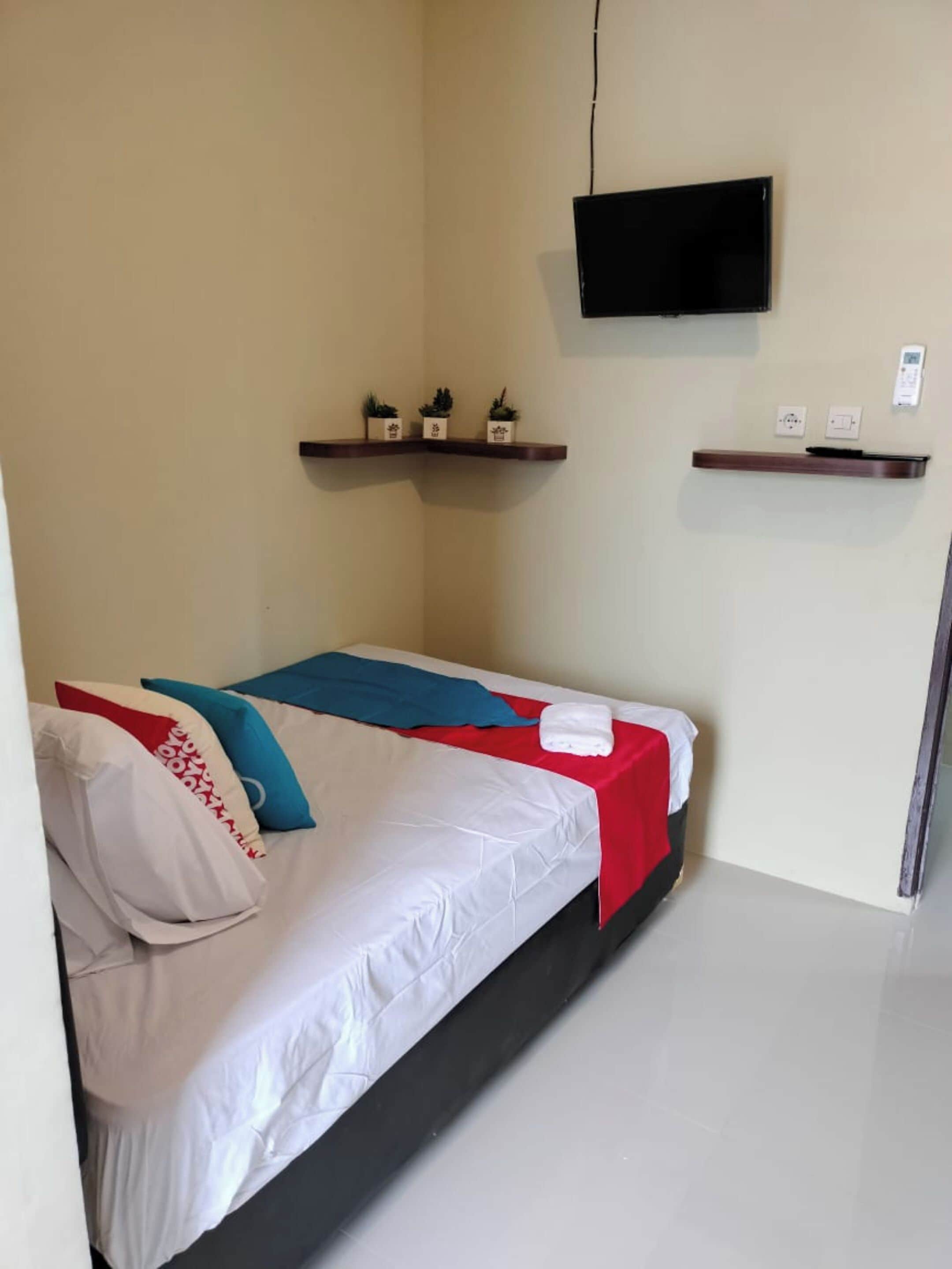 A single bed with white bedding, a red and blue blanket, and a pillow with red and white patterns. There is a flat-screen TV mounted on the wall above the bed.