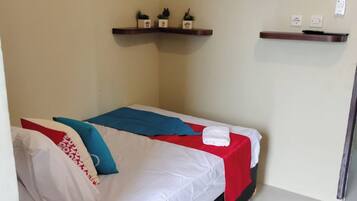 A single bed with white bedding, a red and blue blanket, and a pillow with red and white patterns. There is a flat-screen TV mounted on the wall above the bed.