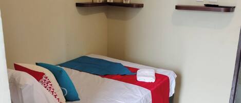 A single bed with white bedding, a red and blue blanket, and a pillow with red and white patterns. There is a flat-screen TV mounted on the wall above the bed.