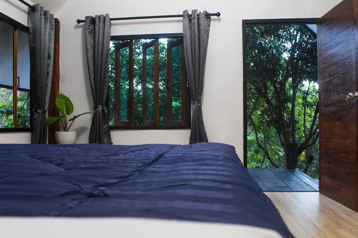 A bedroom with a bed, a window with curtains, a potted plant, and a view of greenery outside.