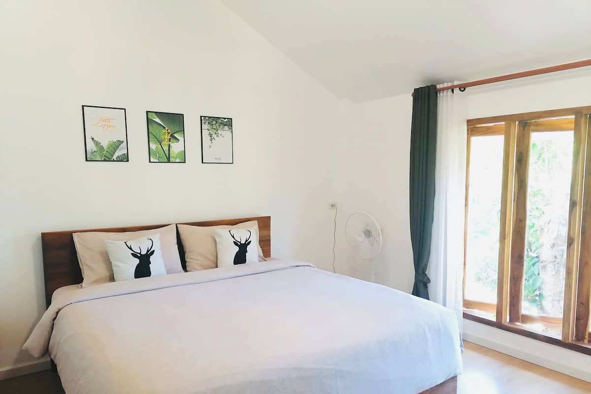 A bedroom with a bed, a wooden headboard, three wall art pieces, a standing fan, and a large window with curtains.