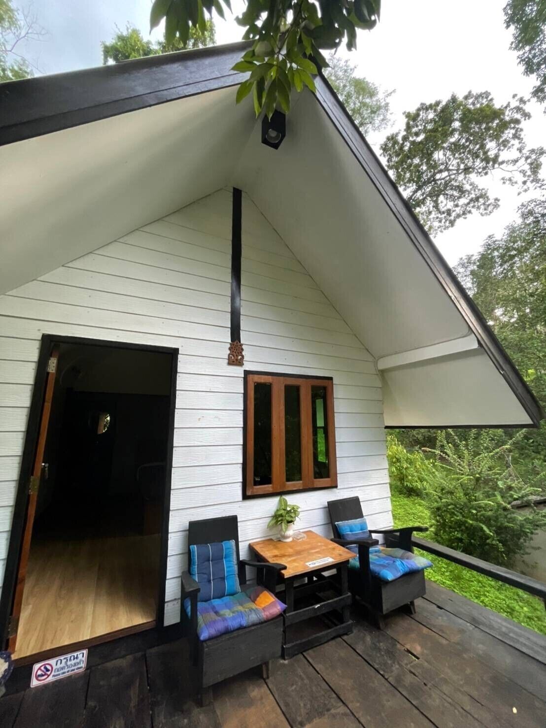 Eine kleine, weiße Holzhütte mit Satteldach, einer Holzterrasse, zwei schwarzen Stühlen mit bunten Kissen und einem kleinen Tisch.