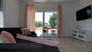 A living room with a sofa, a TV, a small table, and a view of an outdoor area with a pool and greenery. - Luxurious home in Nin (Nin)