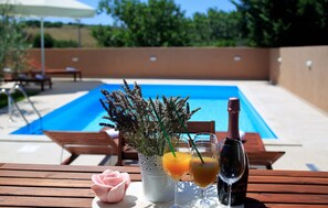 Un entorno junto a la piscina con una mesa de madera, una rosa, un florero con lavanda, dos vasos de jugo de naranja y una botella de vino.