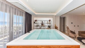 Une piscine intérieure moderne, dotée d’un sol en bois, d’une bibliothèque aux lignes géométriques agrémentée d’objets décoratifs, et offrant une vue sur la mer grâce à de grandes fenêtres.