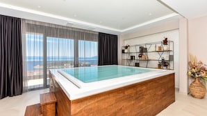 Une piscine intérieure moderne avec une terrasse en bois, de grandes fenêtres dotées de rideaux et une étagère décorative agrémentée de fleurs.