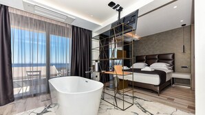 Une chambre d’hôtel moderne dotée d’une grande baignoire, d’un lit avec une tête de lit, d’un téléviseur fixé au mur et d’un balcon avec vue.