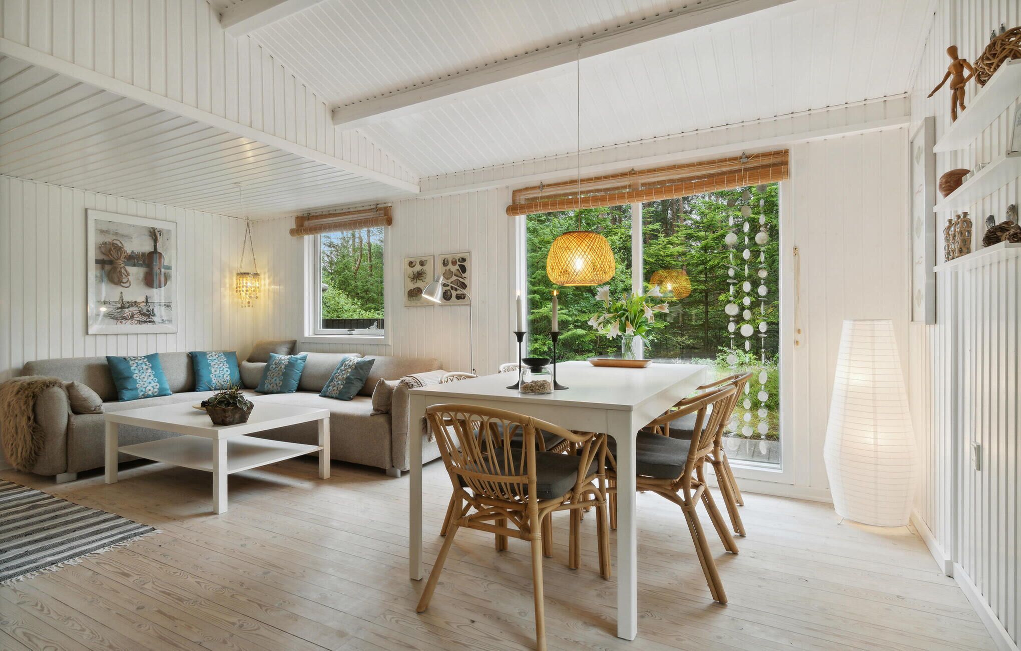 A spacious room with a white ceiling, a dining table with chairs, a sofa with patterned cushions, and a large window with a view of greenery.