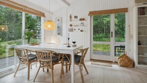 A dining area with a white table and wicker chairs, large windows with a view of greenery, and a shelf with decorative items. - Nice home in ålbæk with WiFi (Ålbæk)