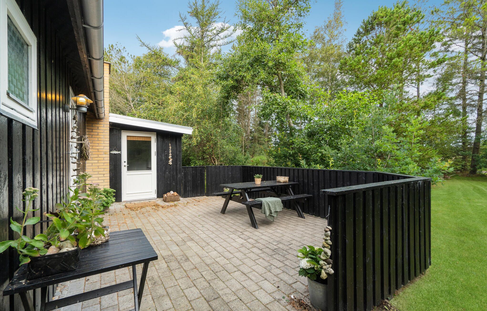 Eine Terrasse mit Holzdeck, einem schwarzen Tisch und einer Bank. Ein kleiner Schuppen mit einer weißen Tür und ein schwarzer Zaun begrenzen den Bereich.