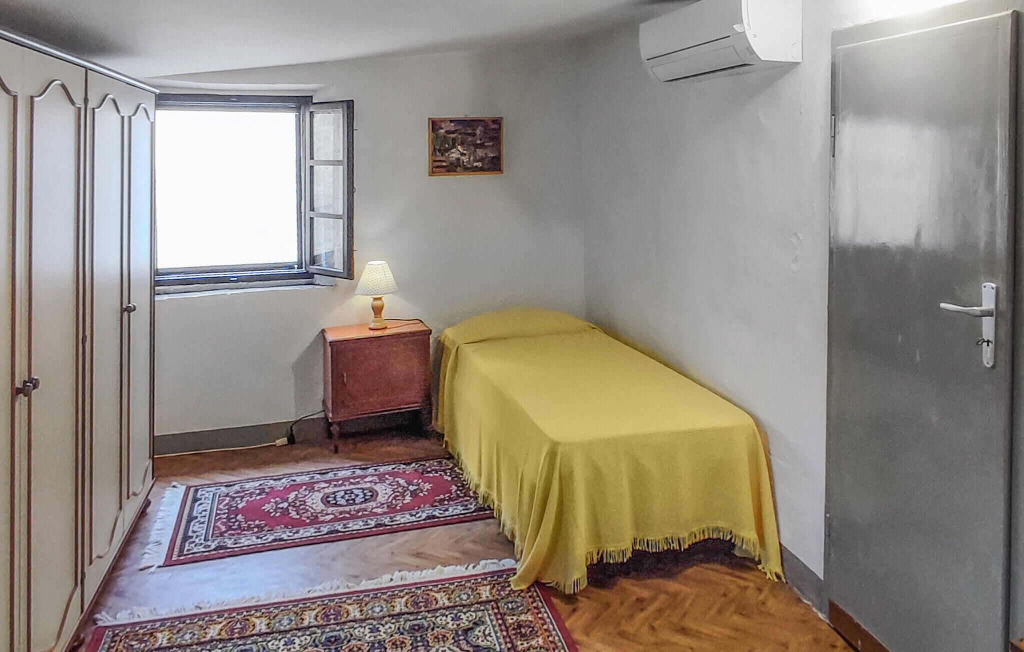Una habitación pequeña con cama, mesita de noche, lámpara, ventana, puerta y aire acondicionado instalado en la pared.