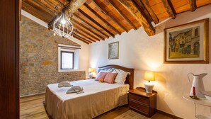 A bedroom with a stone wall, wooden ceiling beams, a bed with a wooden headboard, bedside tables with lamps, and a painting on the wall. - Lovely home in Marliana with WiFi (Marliana)