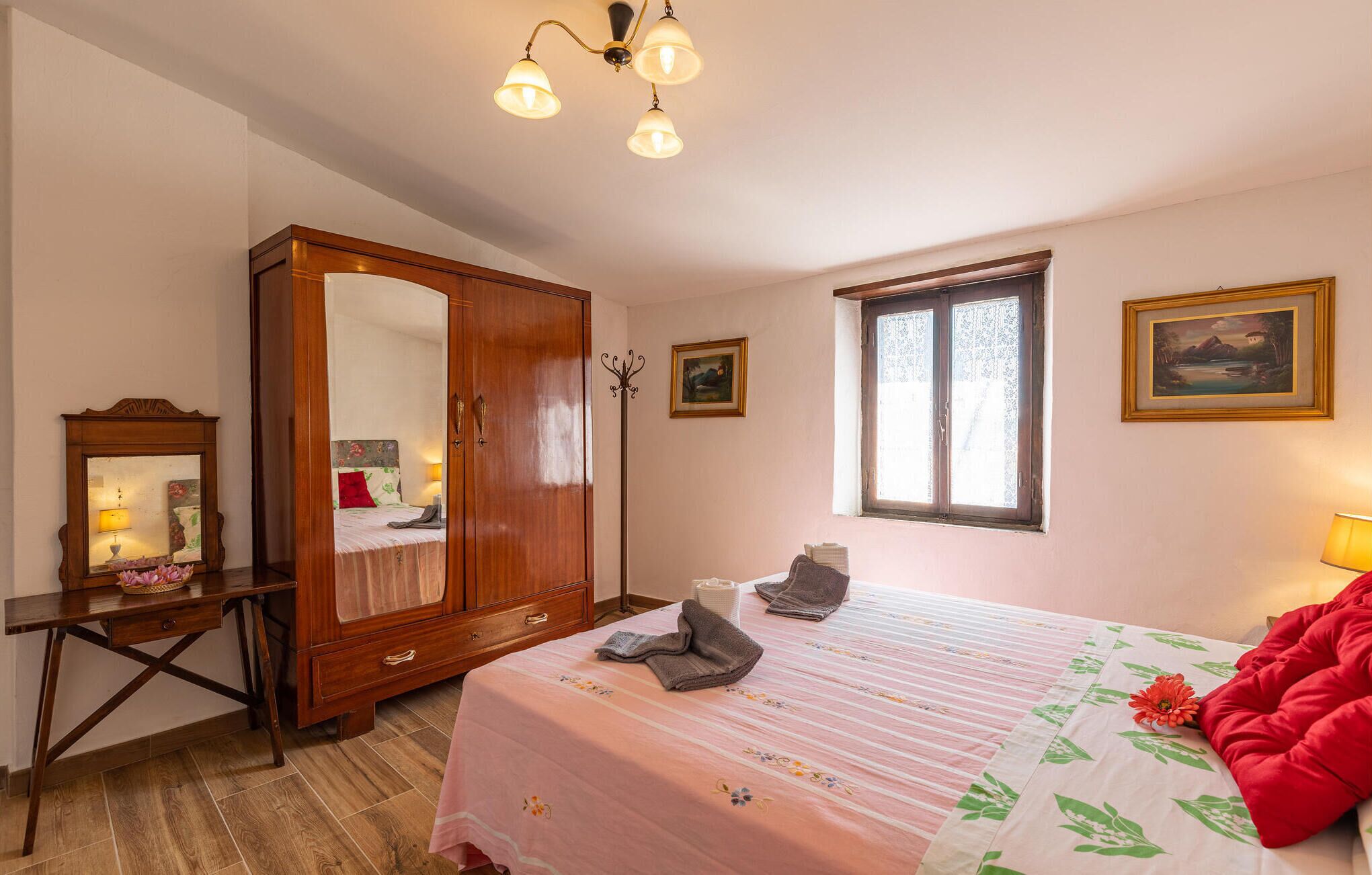 Een slaapkamer met een houten garderobe, een bed met bloemenbeddengoed, een bureau met lamp en een raam met kantgordijnen.
