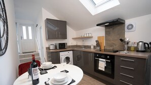 A modern kitchen with a built-in oven, microwave, and washing machine. There is a dining table with a bottle of wine and glasses.
