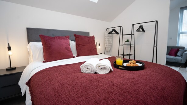 A modern bedroom with a large bed, red bedding, a bedside table with a lamp, a tray with breakfast, and a view of the sky through a skylight.