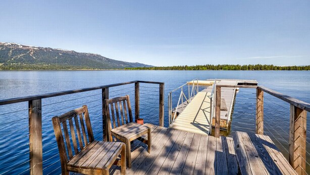 A wooden deck with chairs and a table, overlooking a calm lake with mountains in the background. - Lakeside Cabin With Private Dock & Sandy Beach (Donnelly)