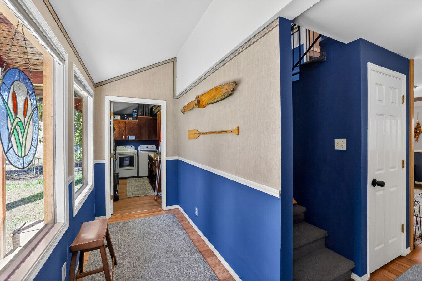 Un pasillo con paredes azules, una puerta de madera, un taburete pequeño y una ventana con un diseño de vitral.