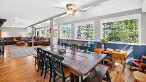 A spacious living area with a wooden dining table, multiple seating options, and large windows offering a view of trees. - Lakeside Cabin With Private Dock & Sandy Beach (Donnelly)